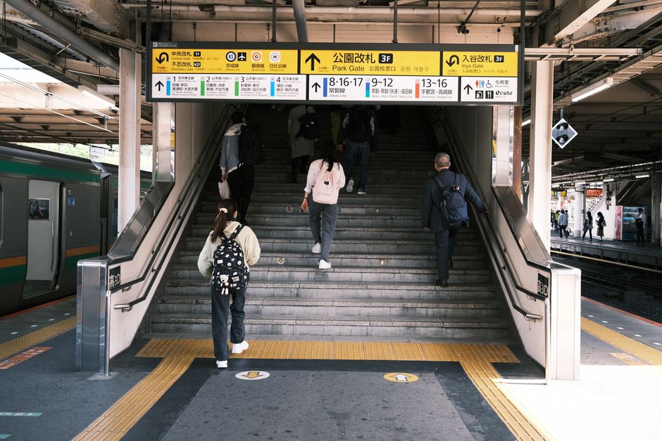 A group of people, including adults and a child, are walking up a staircase inside a train station, preparing for a home relocation or transfer to another part of the station. The staircase, made of grey stone, leads to an upper level with a yellow and white signage board displaying platform information in both English and Chinese, indicating directions for train lines and destinations. Some individuals are carrying backpacks or luggage, suggesting they are passengers en route or arriving. To the left, a train is parked on the platform, partially visible with its green and grey exterior. The station environment features metal structural supports, illuminated signage, and additional passengers on the platform in the background. The scene captures the typical process of packing and moving through a transportation hub, with people engaging in the loading process of their travel or relocation journey, as facilitated by a professional removals service such as Removals Croydon.