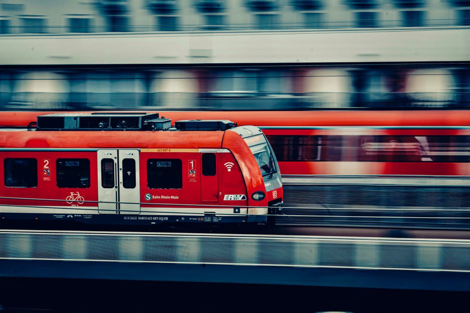 A modern red passenger train traveling at high speed on railway tracks with a blurred background of a station platform. The train has multiple windows, some with reflections, and features emergency and Wi-Fi symbols near the doors. Inside, the train appears to be moving through an urban environment, indicated by the motion blur of the station or cityscape behind it. This image illustrates the speed and efficiency of urban transport, relevant to home relocation or moving services such as those offered by Removals Croydon, especially in the context of quick moves connected to the East Croydon station removals guide.